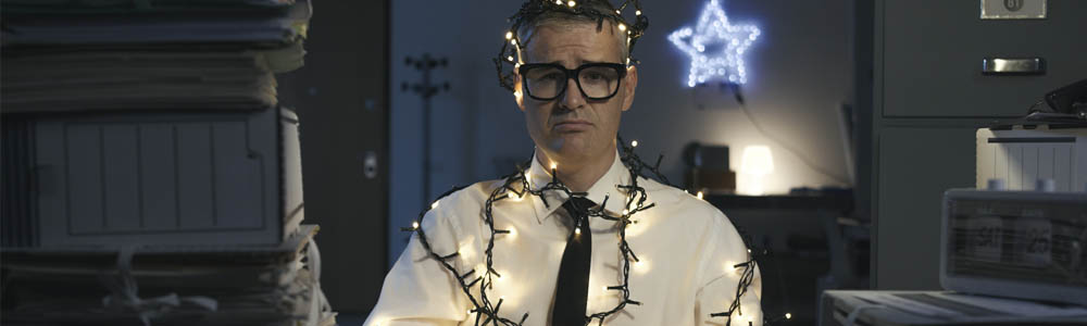 Man in an office wrapped in Christmas lights with work stacked high on his desk, he looks miserable